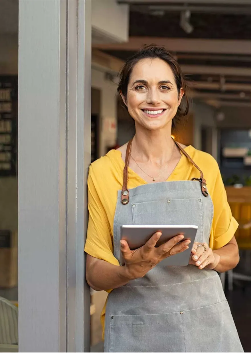 A restaurant owner in an apron holding a tablet in her dining room.